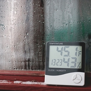 A window with condensation forming, with a hygrometer showing high humidity, that causes crawl space condensation in Ashburn, VA.