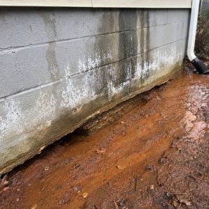 Eroded clay soil at foundation base from poor drainage and downspout discharge causing uneven basement floor in Southbridge, VA.